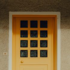 A modern house entrance featuring a bright yellow door with glass panels, casting afternoon shadows.