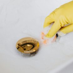 From above of crop anonymous person in yellow rubber protective glove using sponge and detergent for washing white sink in bathroom