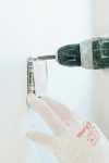 Gloved hands using a drill to install a power outlet on a wall, focusing on precision and safety.