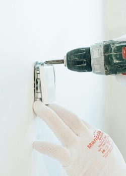Gloved hands using a drill to install a power outlet on a wall, focusing on precision and safety.