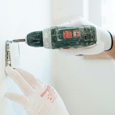 Gloved hands using a drill to install a power outlet on a wall, focusing on precision and safety.